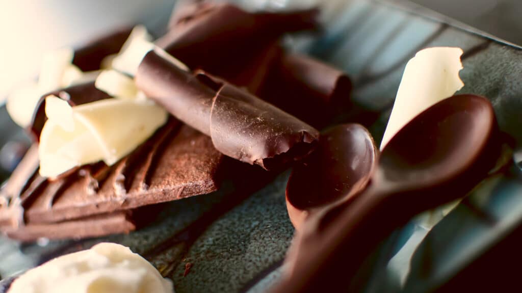 chocolate square cookie draped in chocolate and fudge sauce, with white chocolate curls and dark chocolate shavings and chocolate spoons for coffee or dessert